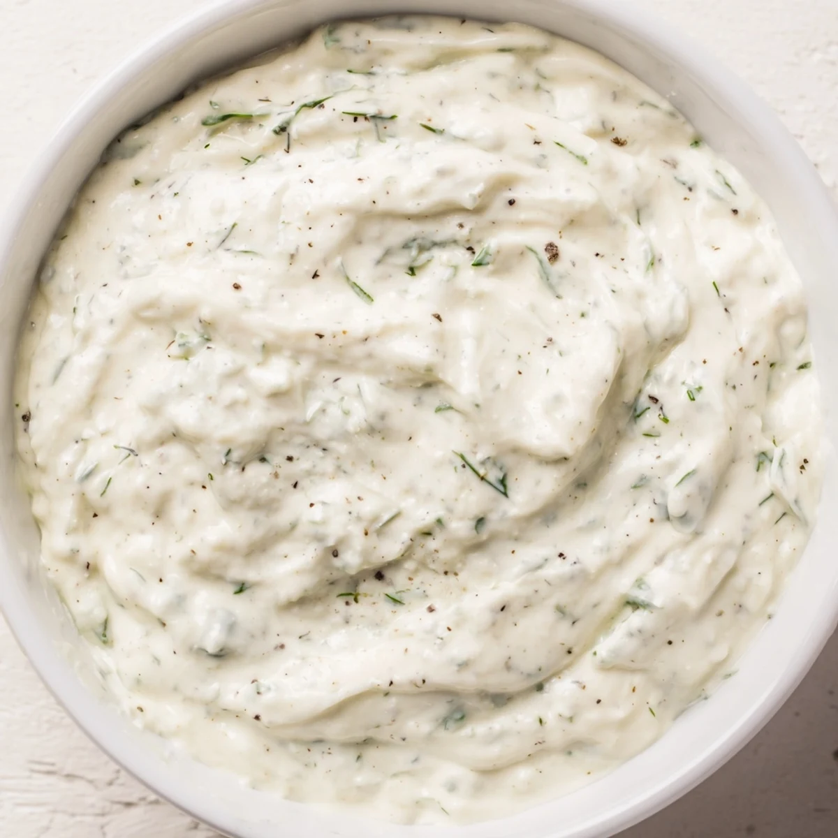 A whisk dips into a bowl of Creamy Salad Dressing with Herbs, showcasing its thick, herby texture against a bright kitchen counter.