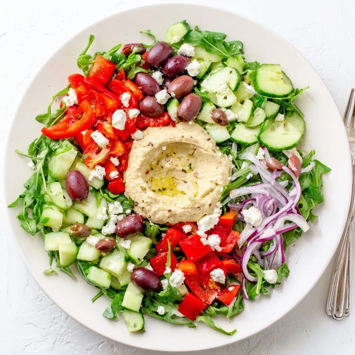 A close-up of Mediterranean Salad with Hummus reveals crumbled feta, Kalamata olives, and a generous dollop of hummus on a colorful bed of greens.