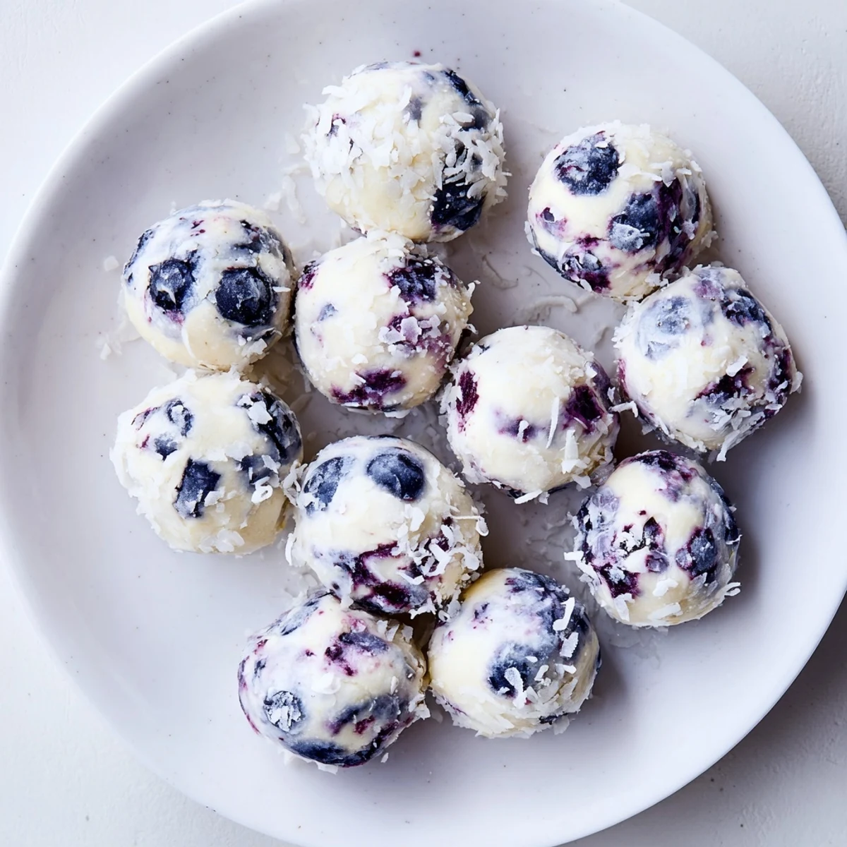 Golden blueberry cheesecake protein bites arranged on a white plate, topped with fresh blueberries and coconut flakes