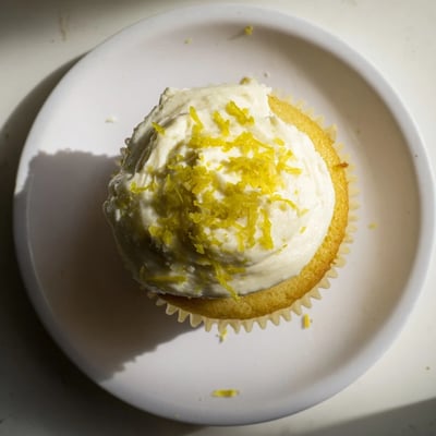 A close-up of frosted Limoncello Cupcakes with lemon zest garnish beside a glass of golden liqueur.  